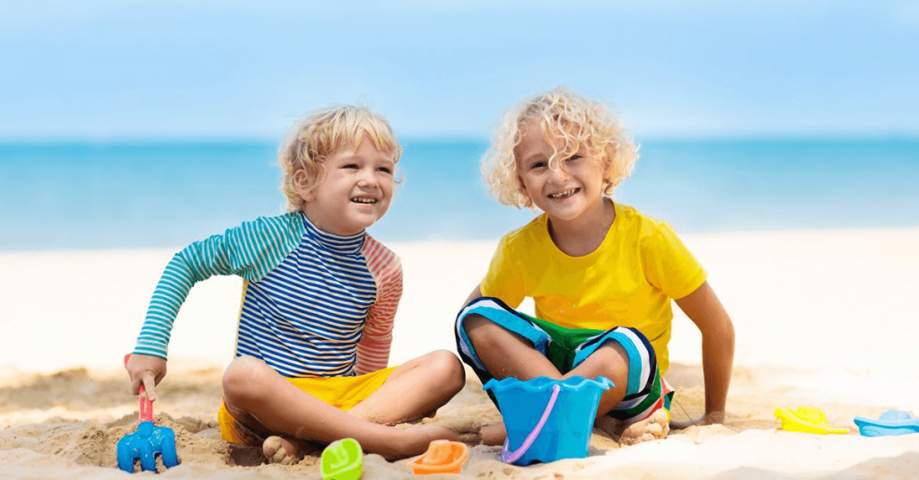 young boy with long sleeved multicolored rashguard shirt and yellow swim trunks next to young boy with yellow tshirt and mutlicolored swim trunks playing in the sand at a beach