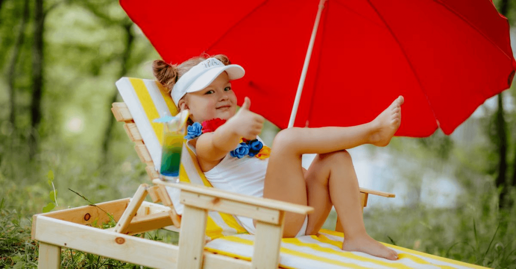 smiling young girl in white bathing suit and white visor laying on yellow and white striped beach chair under a red umbrella with thumbs up to camera