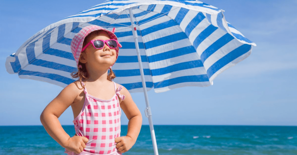 young girl in pink checked bathing suit with pink sunglasses and pink checked hat standing with hands on hips looking toward the sun with water and blue and white striped beach umbrella behind her