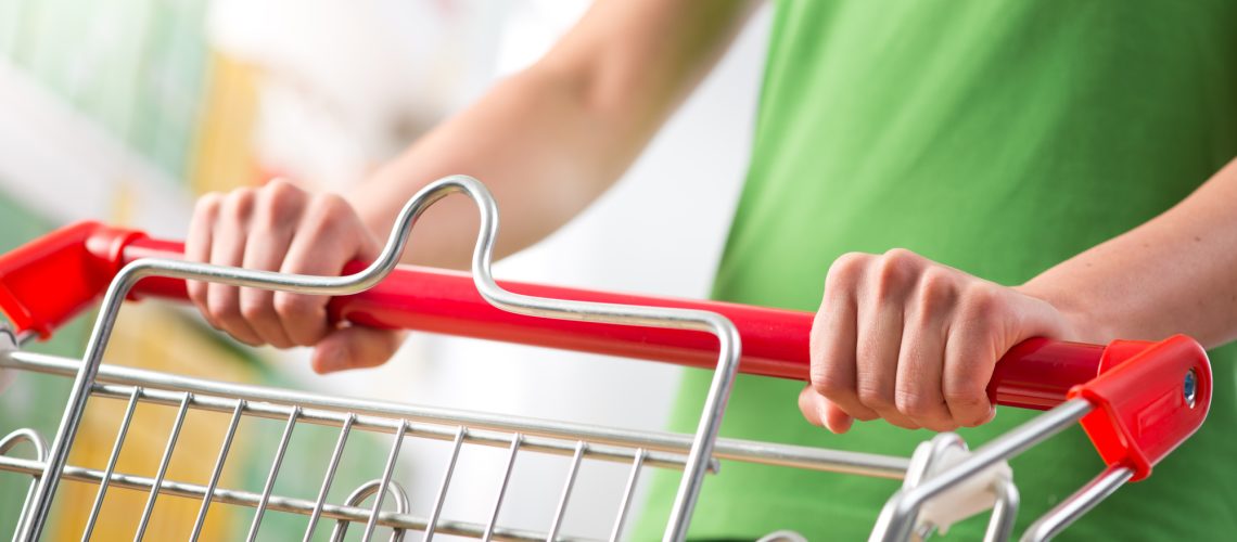Woman in green t-shirt pushing a shopping cart at store with shelves on background.